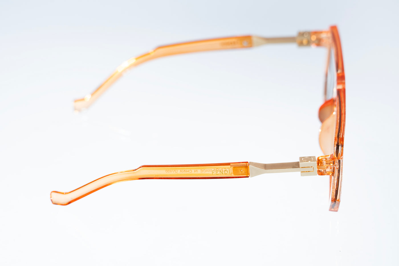 A pair of translucent orange acetate sunglasses with gold metal hinges, shown from a side profile view against a white background.
