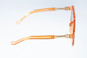 A pair of translucent orange acetate sunglasses with gold metal hinges, shown from a side profile view against a white background.
